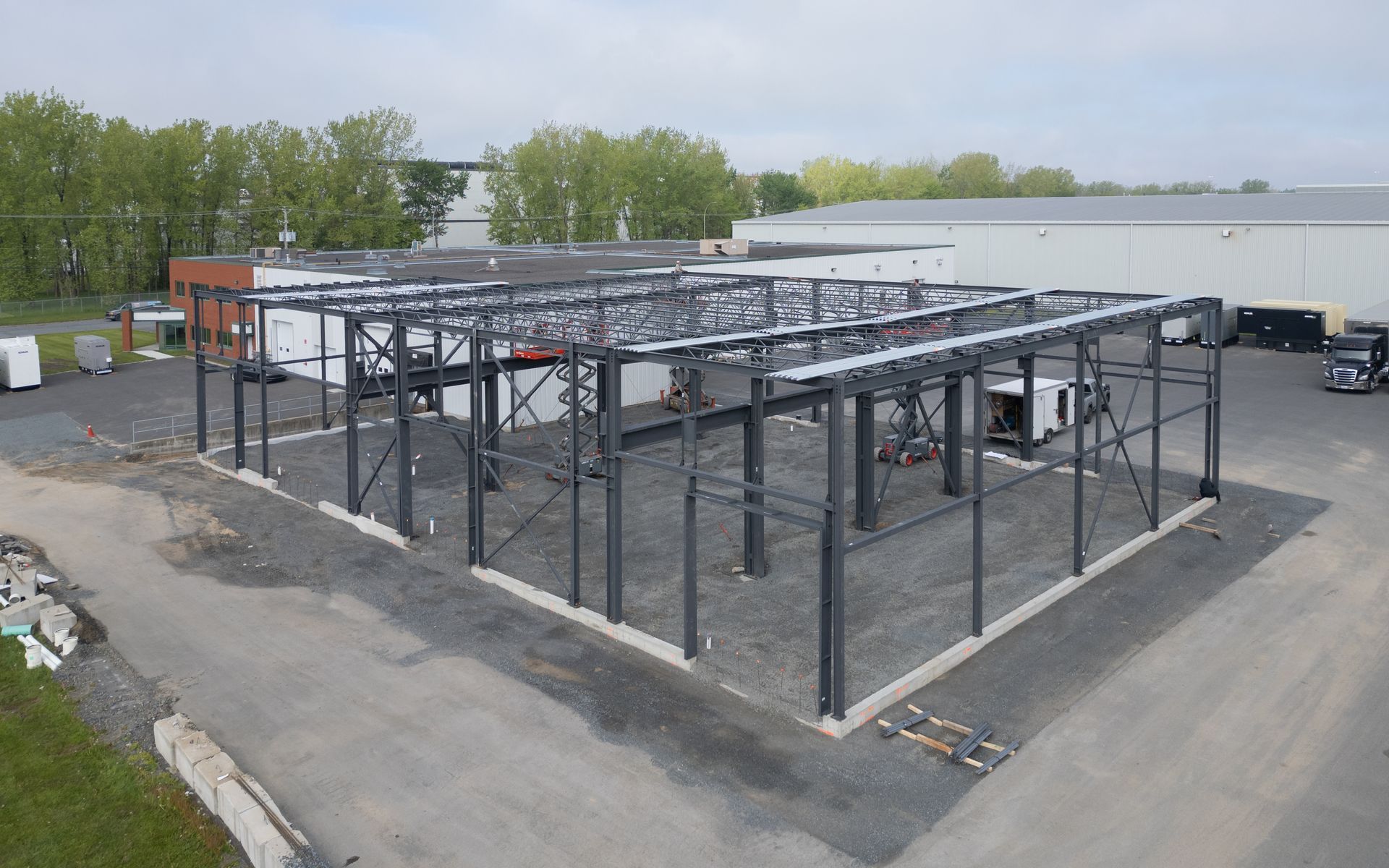Ossature métallique d'un bâtiment en construction, acier gris, fondations en gravier, autres bâtiments en arrière-plan.