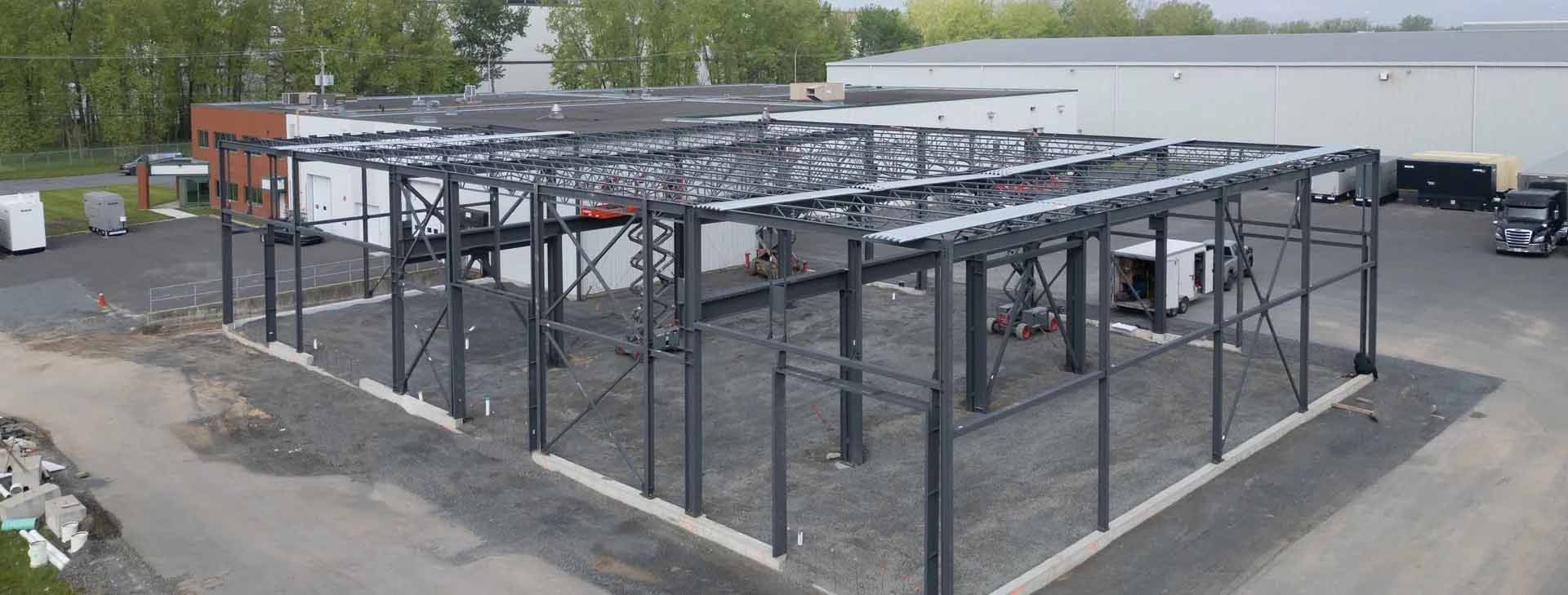 Bâtiment à ossature métallique en construction sur un terrain gravillonné, avec des fermes de toit inachevées et des camions alentour.