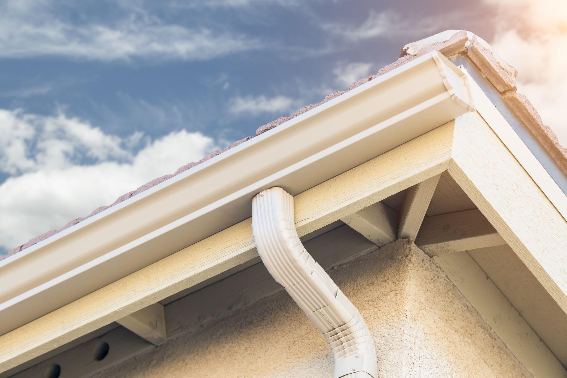 White vinyl rain gutter and downspout on home roof corner under blue sky, exterior drainage detail.