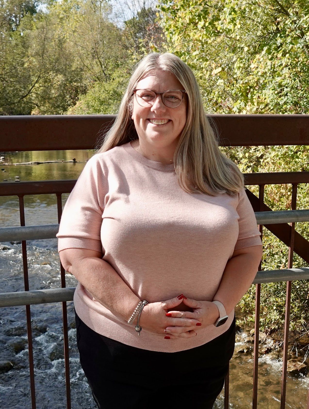 Woman in pink top and glasses smiles on a bridge over a river.