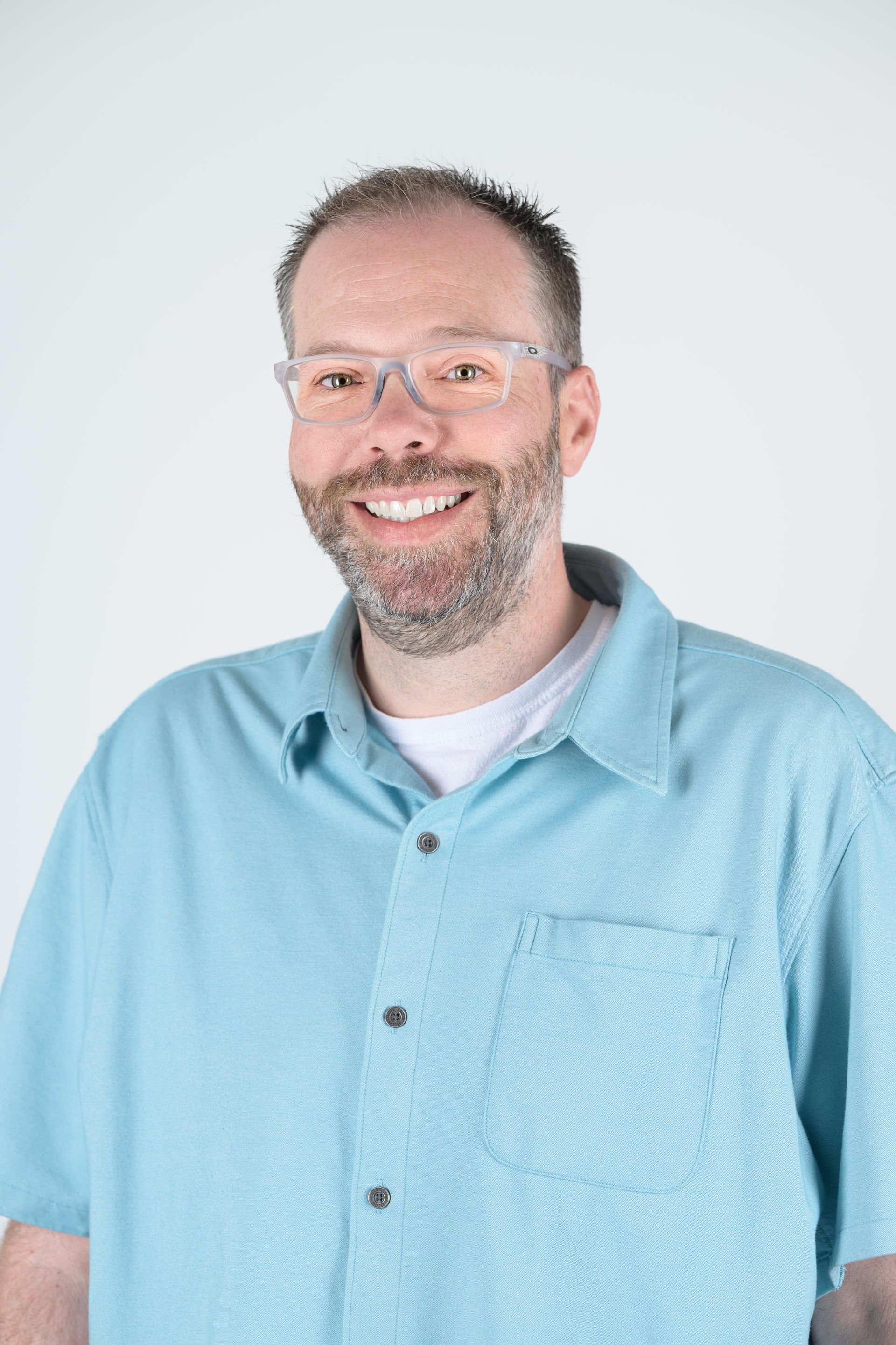 Man in glasses smiles, wearing a light blue button-up shirt and a white undershirt, against a white backdrop.