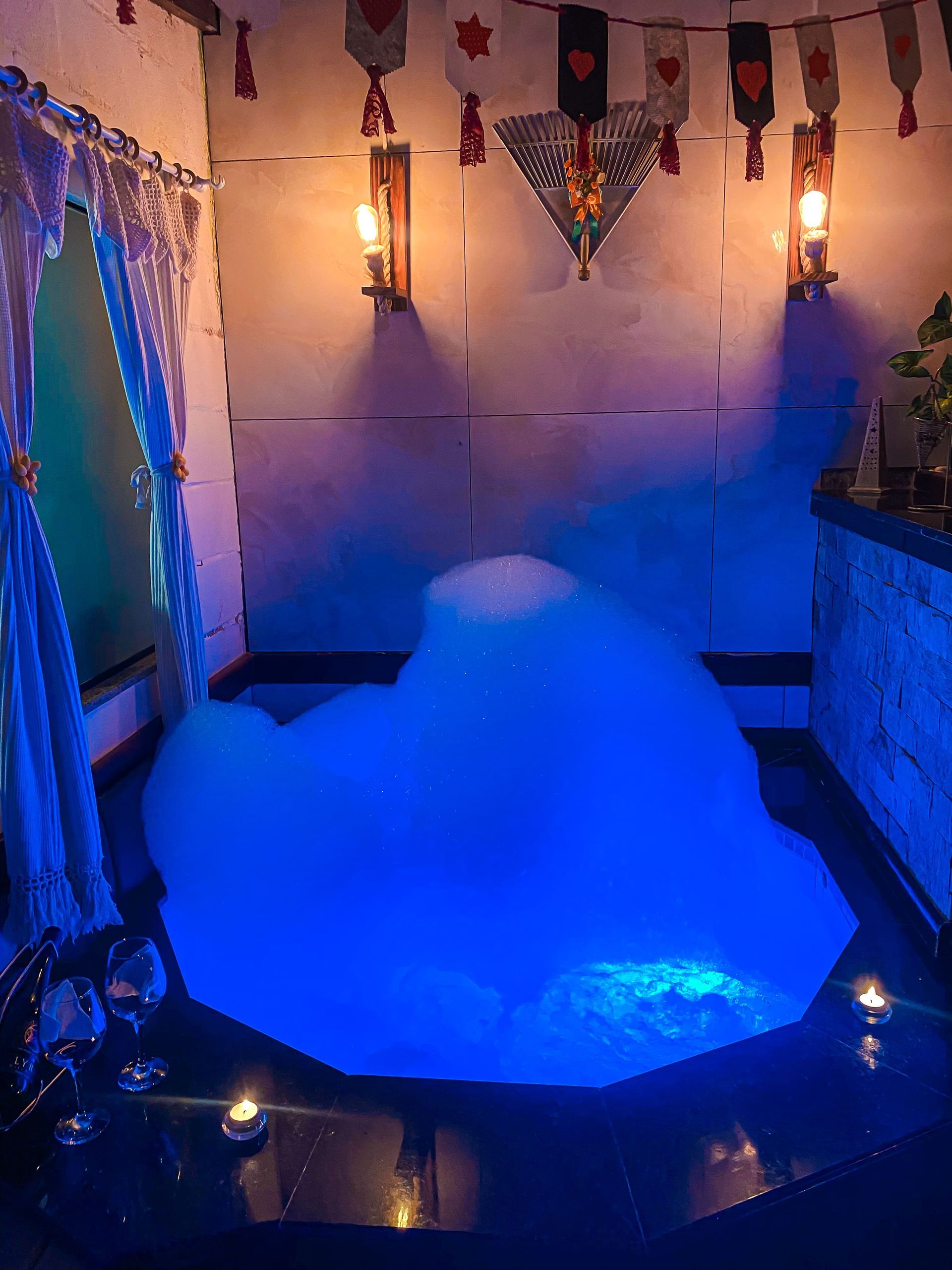 Um quarto romântico com iluminação suave apresenta uma banheira redonda cheia de espuma de banho azul brilhante, iluminada por luminárias de parede e velas.