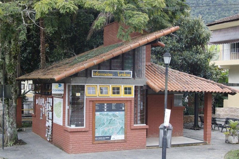 Um quiosque de informações turísticas de tijolos vermelhos com telhado inclinado de telhas e um mapa em exposição, situado sob árvores em um parque.