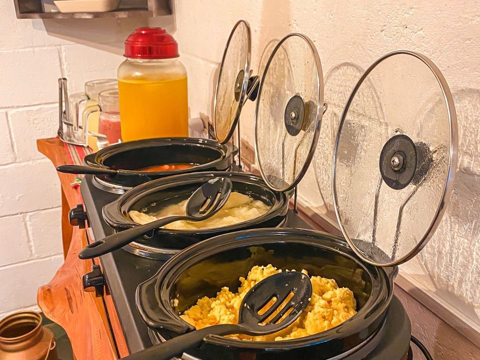 Uma estação de buffet de café da manhã com três panelas elétricas de cozimento lento cheias de comida quente, com as tampas abertas, e um dispensador de suco.