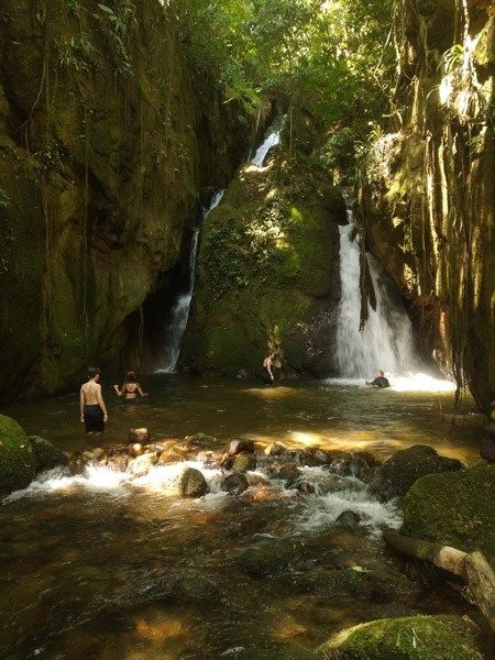 Uma pequena cachoeira sombreada na floresta, que deságua em uma piscina onde as pessoas caminham e nadam entre rochas e vegetação exuberante.