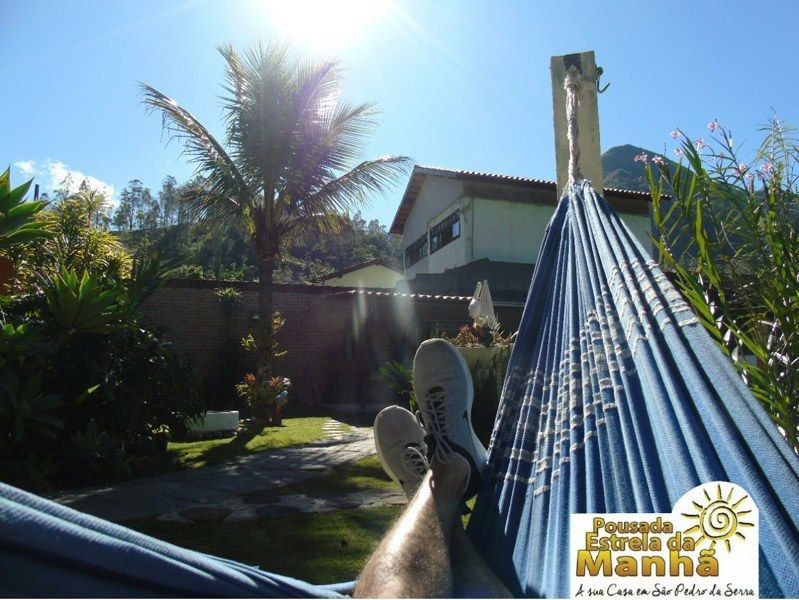 Perspectiva em primeira pessoa de pernas descansando em uma rede azul em um jardim ensolarado com um prédio e uma palmeira ao fundo.