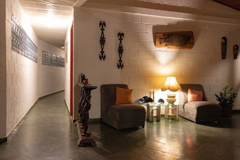 Um corredor pouco iluminado com duas cadeiras, um abajur sobre uma mesa lateral e obras de arte decorativas em uma parede de tijolos brancos.