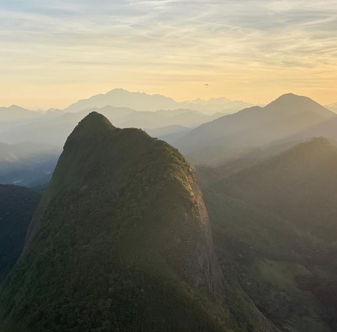 Um pico de montanha iluminado pelo sol se destaca em primeiro plano, com camadas de cadeias montanhosas envoltas em névoa e bruma que se estendem ao longe.