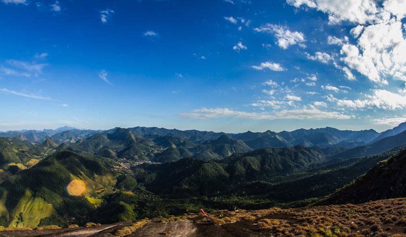 Uma vista panorâmica de exuberantes cadeias de montanhas verdes sob um céu azul brilhante com nuvens brancas e suaves.
