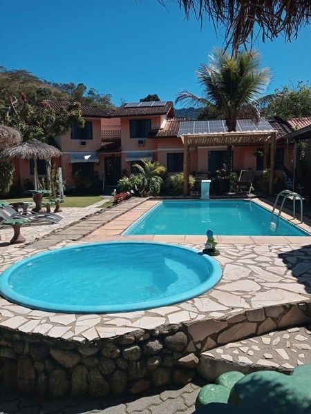Uma área de resort com uma banheira de hidromassagem circular azul em primeiro plano, uma piscina retangular e um edifício de dois andares.