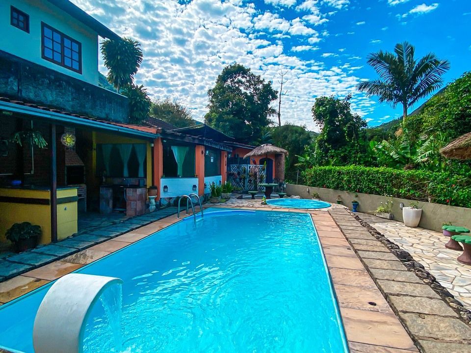 Piscina azul brilhante em um pátio ao lado de uma casa multicolorida com plantas verdes e um céu ensolarado.