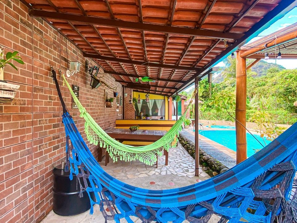 Uma rede verde brilhante está pendurada em um pátio coberto de tijolos ao lado de uma piscina, com árvores exuberantes ao fundo.