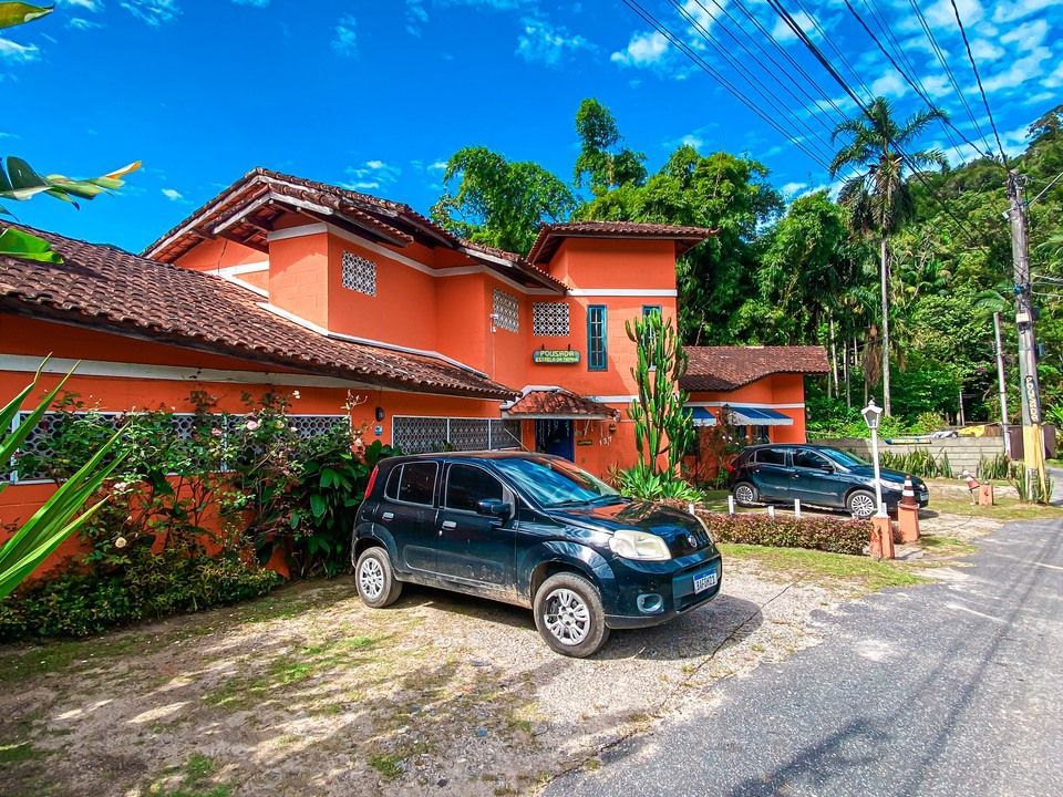 Um carro preto estacionado em frente a uma casa laranja vibrante com telhado de telhas, cercada por árvores verdejantes e um céu azul.