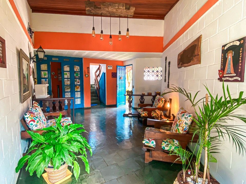 Uma sala de estar interna colorida com poltronas estampadas, plantas exuberantes em vasos, um lustre de madeira no teto e detalhes em laranja vibrante.