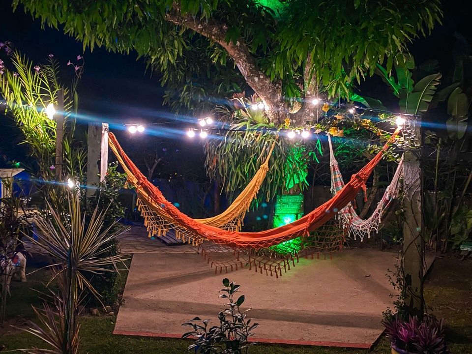 Três redes coloridas estão penduradas sob uma grande árvore decorada com luzes de corda em um jardim à noite.