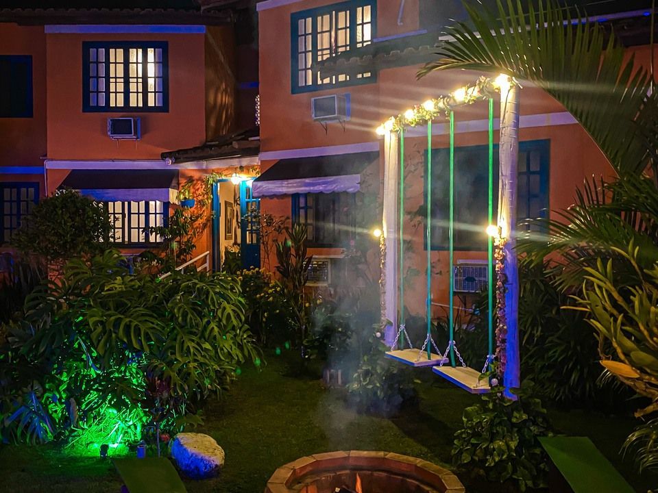 À noite, um aconchegante pátio externo conta com uma fogueira iluminada, dois balanços de madeira e construções alaranjadas com iluminação quente.