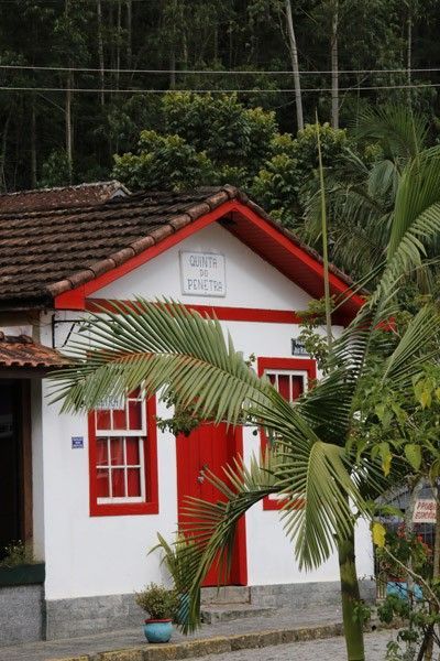Uma casa branca com detalhes em vermelho vivo, uma porta vermelha e um telhado de telhas, parcialmente encoberta por uma palmeira.