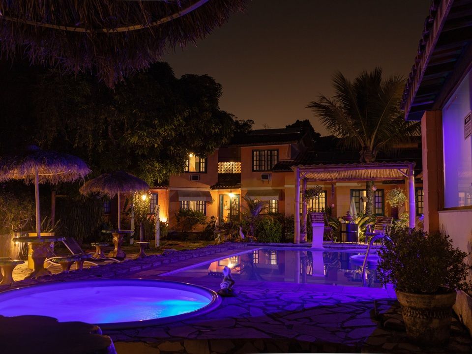 À noite, uma piscina e uma banheira de hidromassagem são iluminadas por uma vibrante luz roxa em frente a um edifício rústico de vários andares.