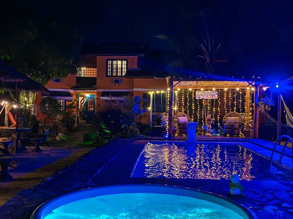 Uma piscina e uma banheira de hidromassagem em uma villa iluminada à noite, com luzes de corda em tons quentes e iluminação subaquática azul na piscina.