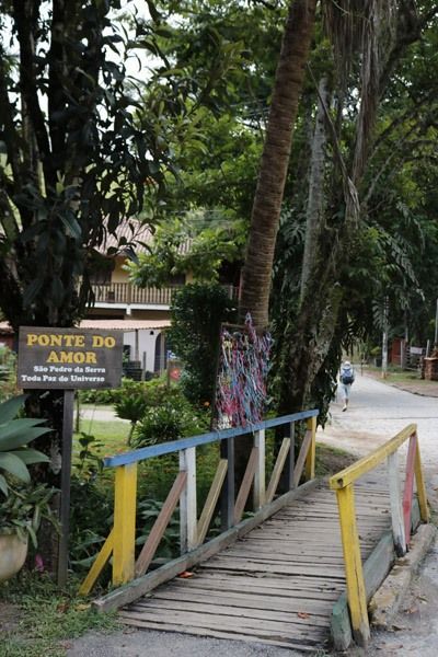 Uma pequena ponte de madeira com grades amarelas e azuis, com uma placa que diz "Ponte do Amor", em um parque.