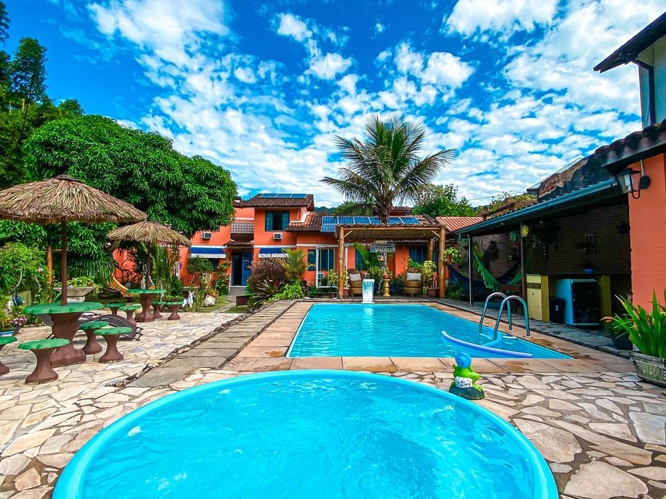 Uma piscina retangular e uma banheira de hidromassagem redonda em uma área externa com palmeiras, espreguiçadeiras e construções laranjas.