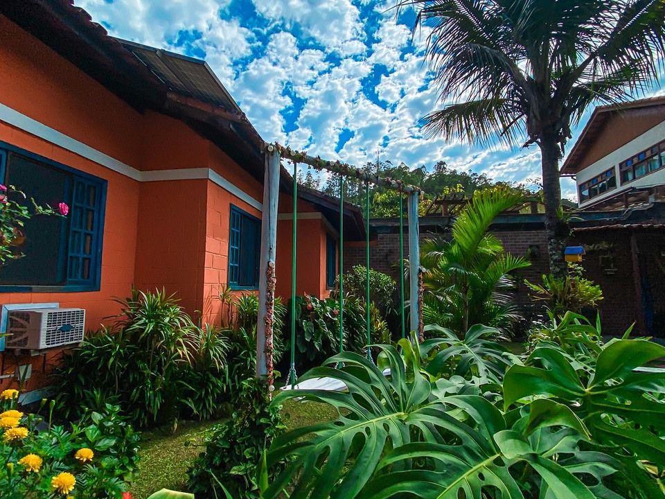 Uma casa laranja vibrante com janelas de moldura azul fica perto de plantas verdejantes e um balanço de madeira sob um céu ensolarado.