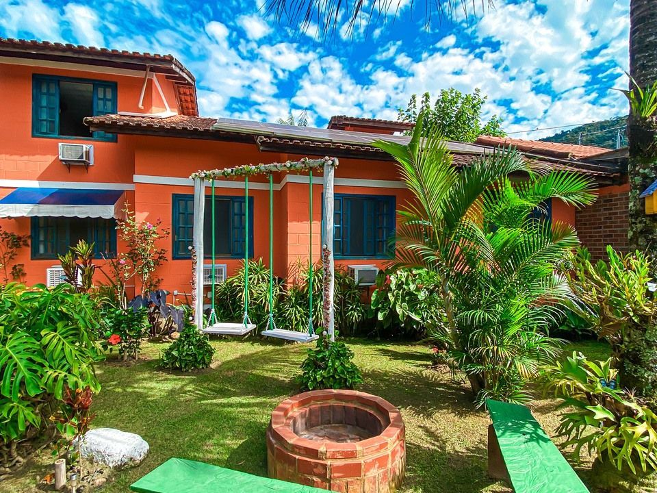 Uma casa laranja vibrante com um quintal que inclui uma lareira de tijolos, bancos verdes e um balanço, sob um céu azul nublado.