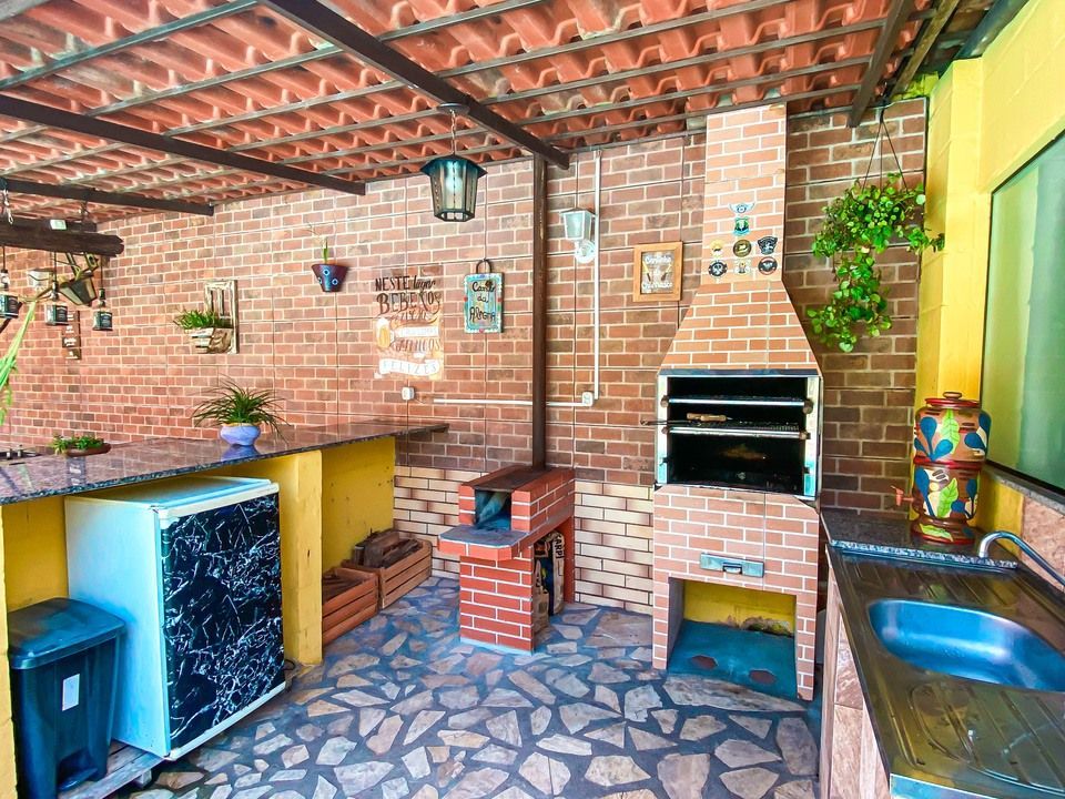 Pátio externo com churrasqueira de tijolos, piso de pedra, pia de metal, frigobar e plantas suspensas sob um telhado de telhas.