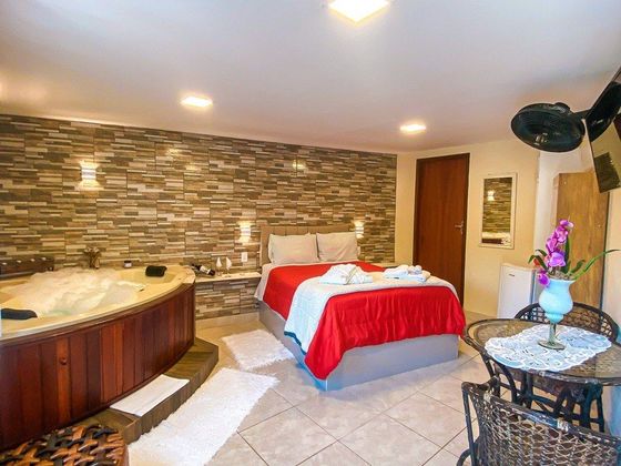 Um quarto de hotel com uma banheira de hidromassagem de madeira, uma cama com roupa de cama vermelha, uma parede de pedra texturizada e uma pequena mesa redonda com cadeiras.