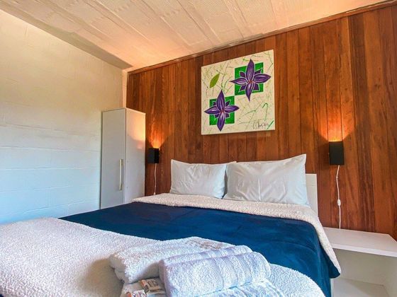 Uma cama impecavelmente arrumada com um cobertor azul e travesseiros brancos encostada em uma parede de madeira com uma obra de arte floral.
