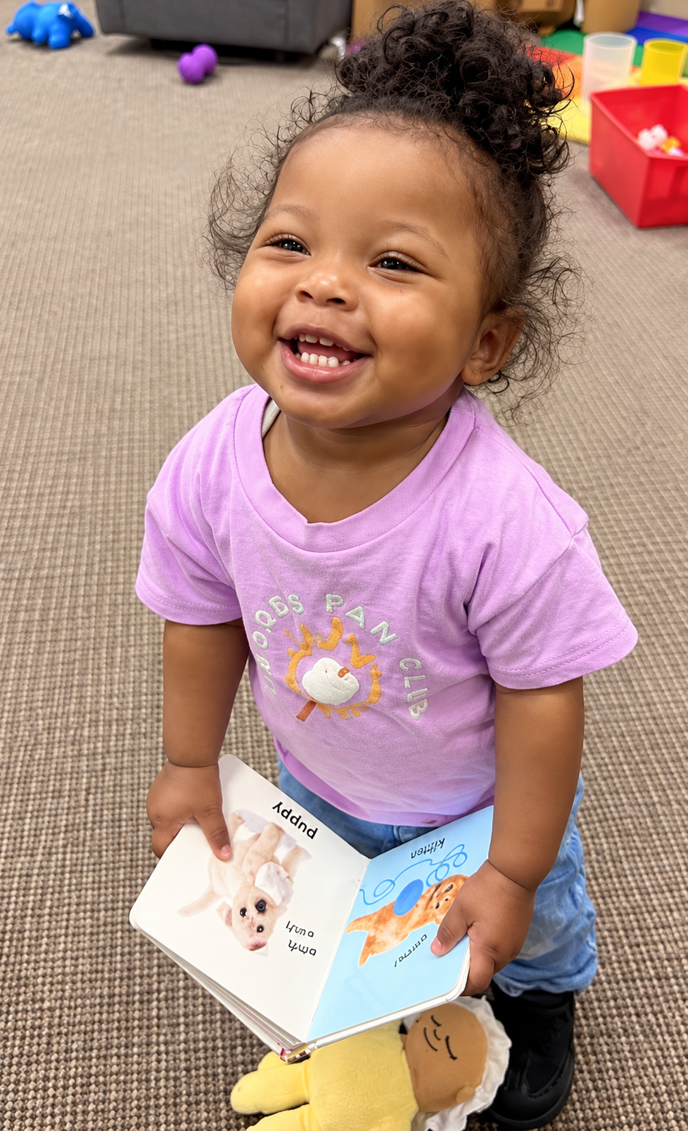 Smiling toddler holding an open book in a playroom with toys on the floor
