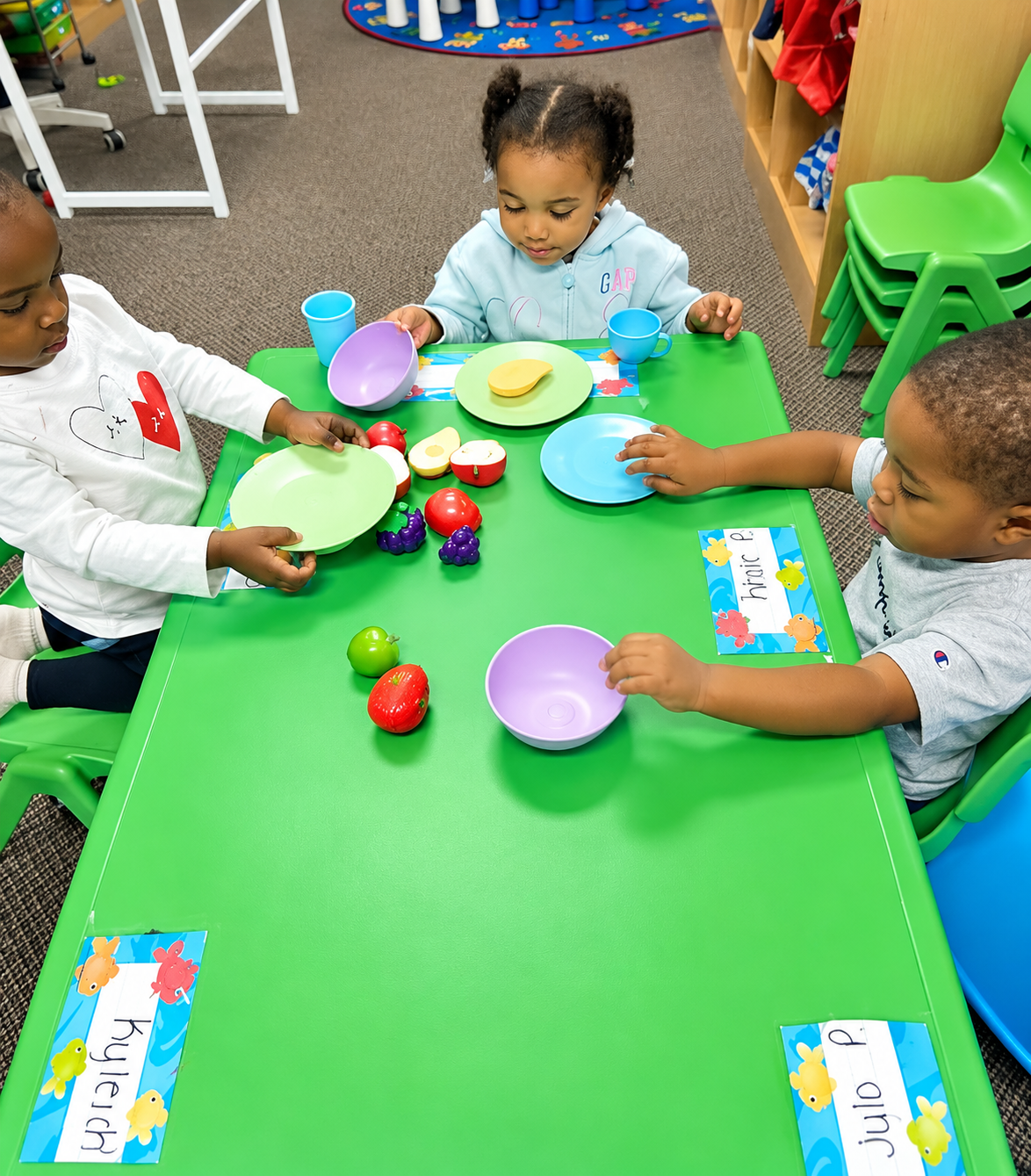Children playing with toy dishes at a green table in a classroom.
