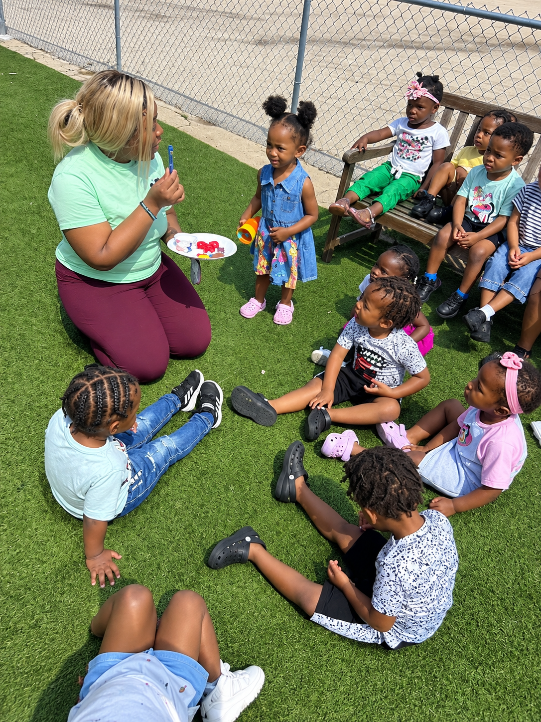 Teacher reading to a small group of children seated on grass outdoors near a fence.