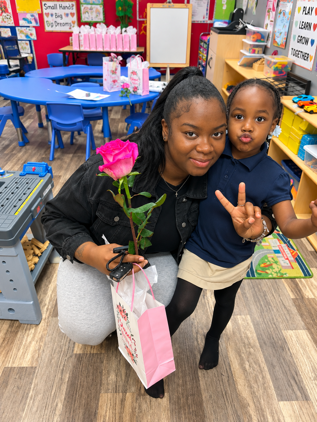 Woman and child posing in a colorful classroom, holding a pink flower and gift bag, both smiling at the camera