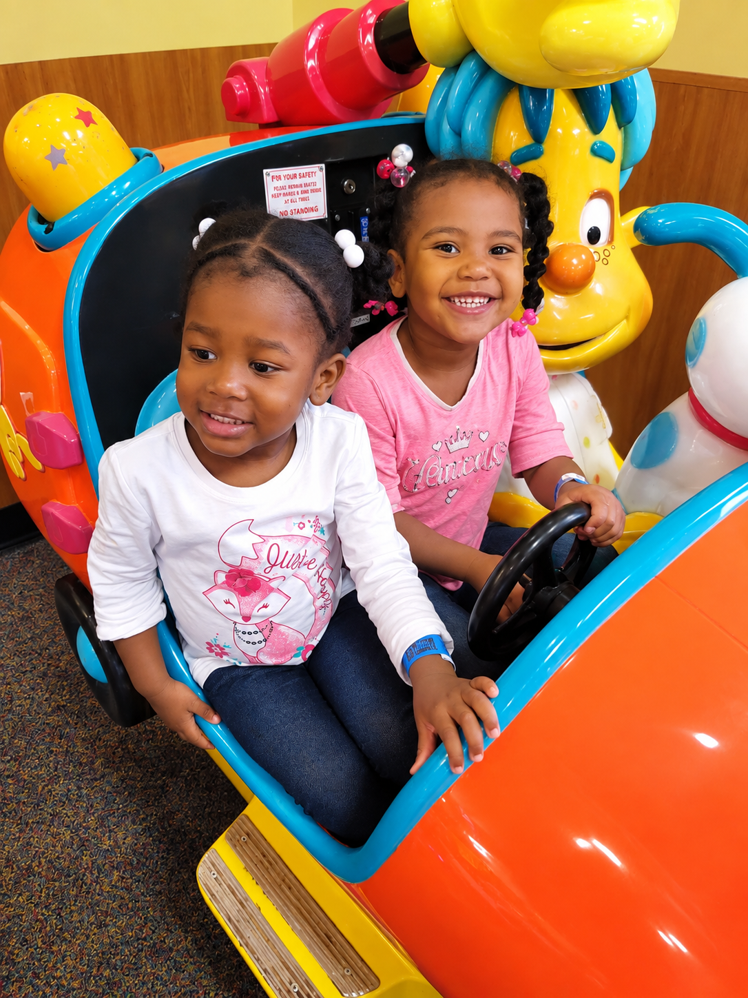 Two children smiling in a colorful amusement ride, one in a white shirt and one in pink.