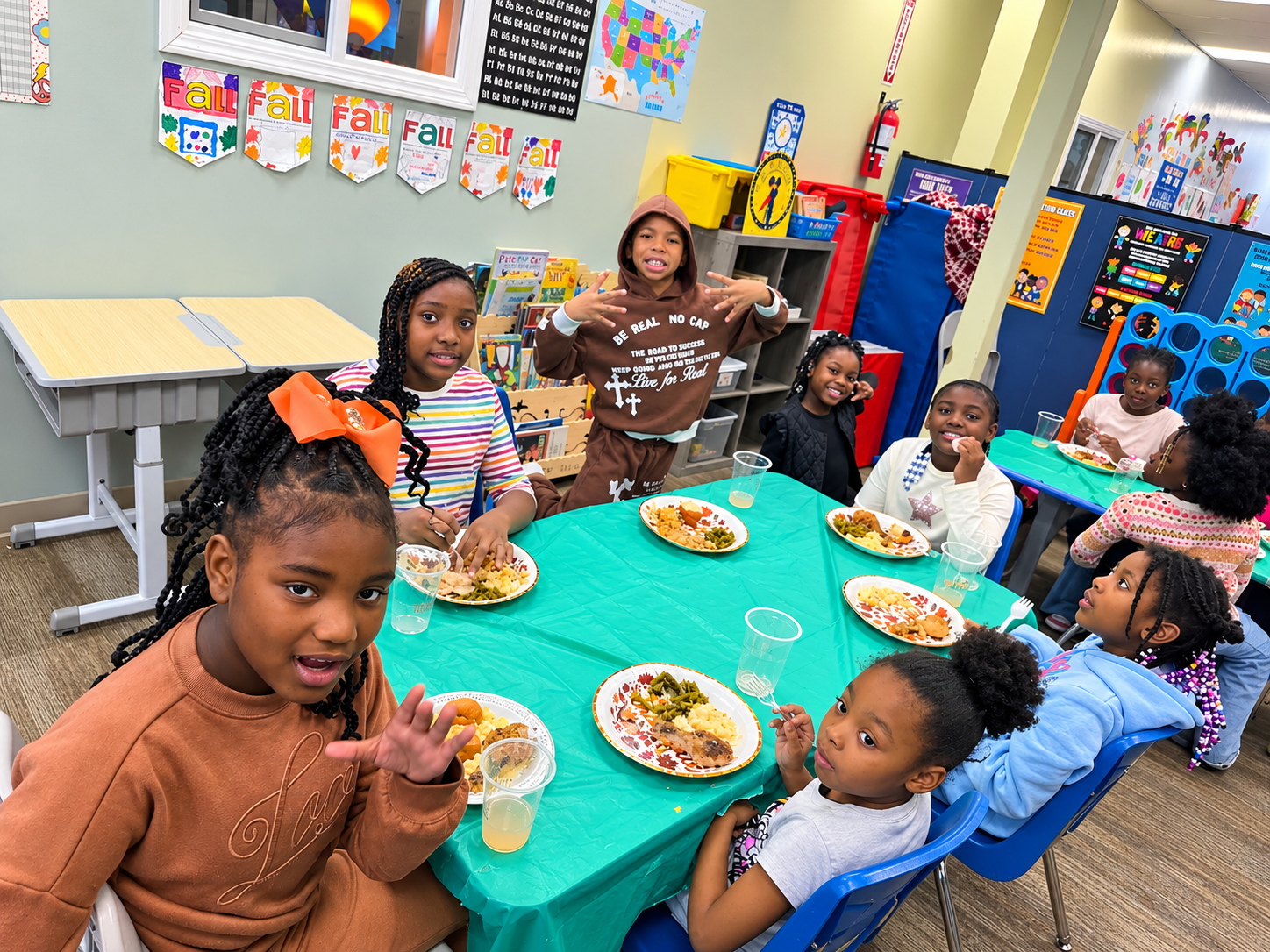 Children seated around a teal table at a colorful classroom party, eating plates of food and smiling.