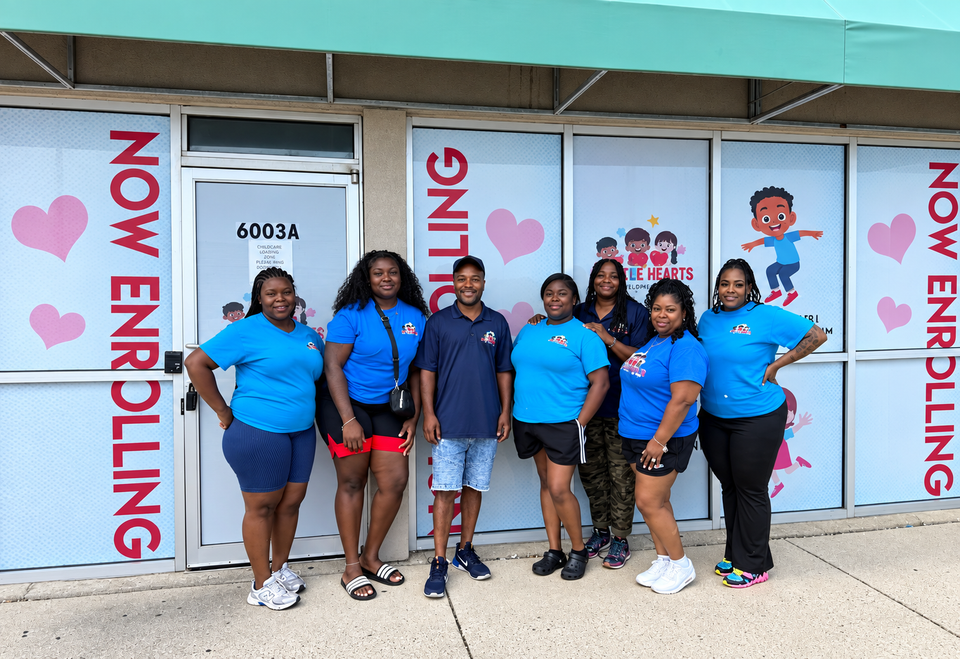Group in blue shirts posing outside a storefront with “NOW ENROLLING” signs and heart decorations.