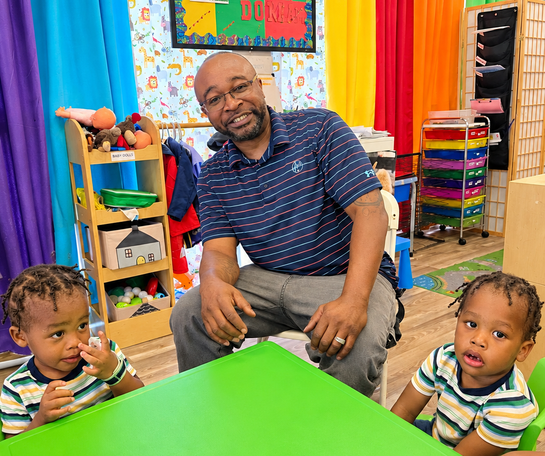 Man sitting with two children at a bright green table in a colorful classroom.