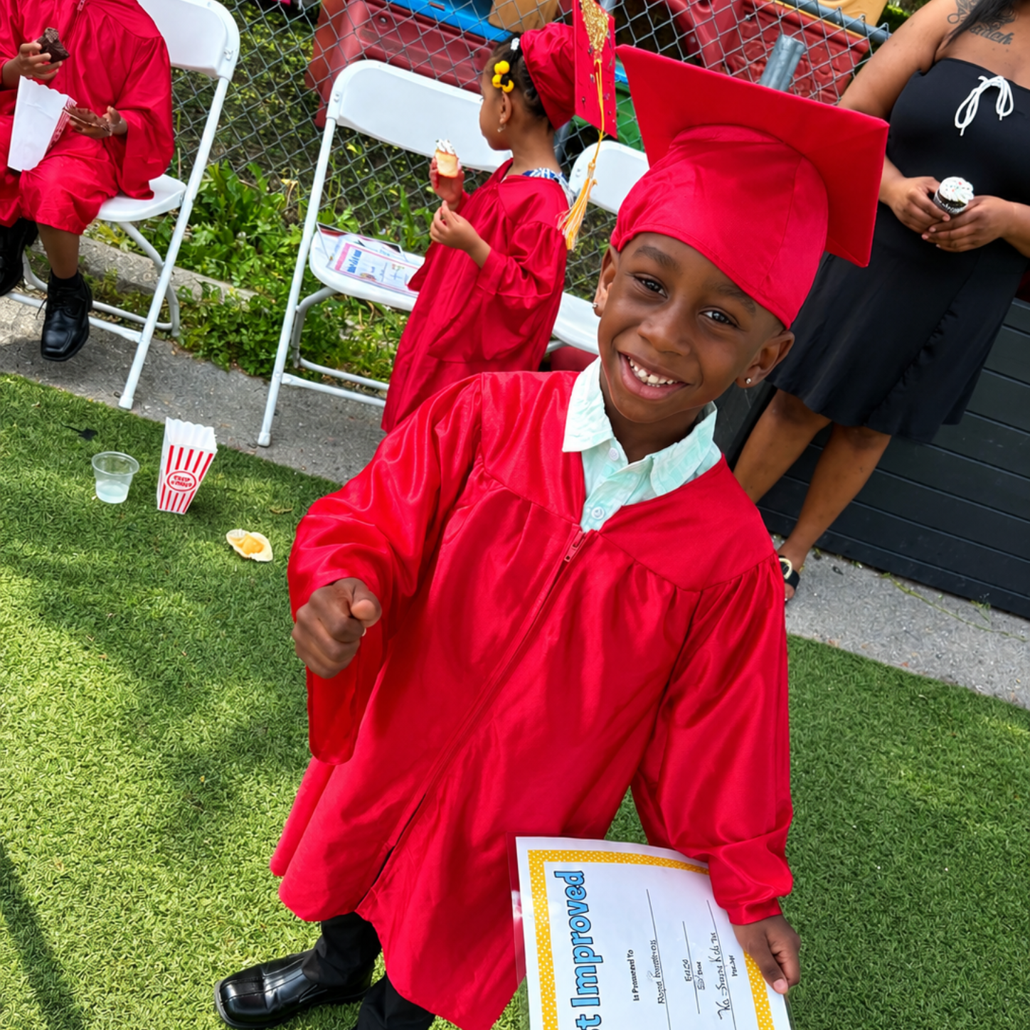 Child in red graduation gown and cap smiling outdoors, holding a certificate.