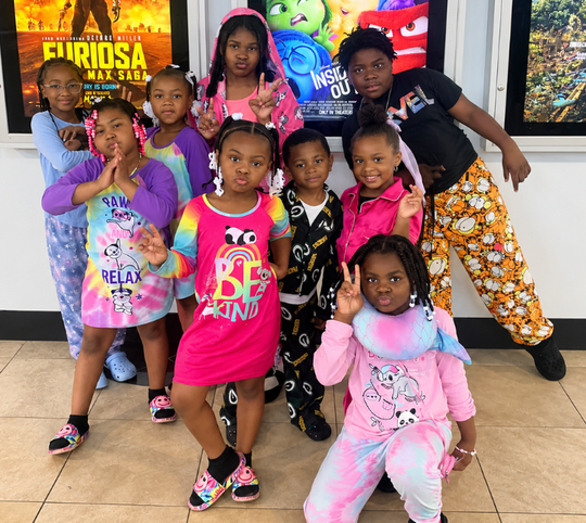 Eight children posing together indoors in colorful outfits in front of a movie poster display.
