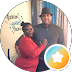 Two people posing indoors; one in red, one in dark clothing, with a circular star badge overlay.