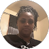 A person in a black shirt with braided hair taking a selfie indoors, with a beige wall behind them.