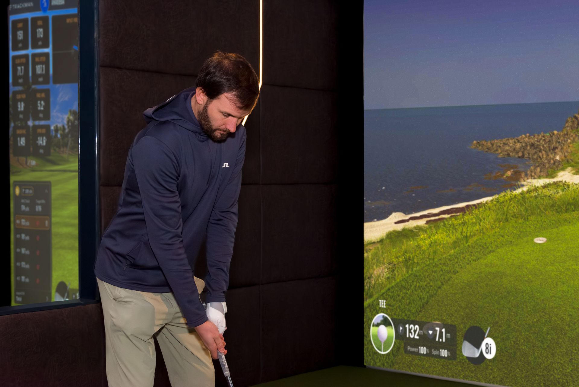 Golfer preparing a swing indoors beside a large golf simulator screen showing a coastal course