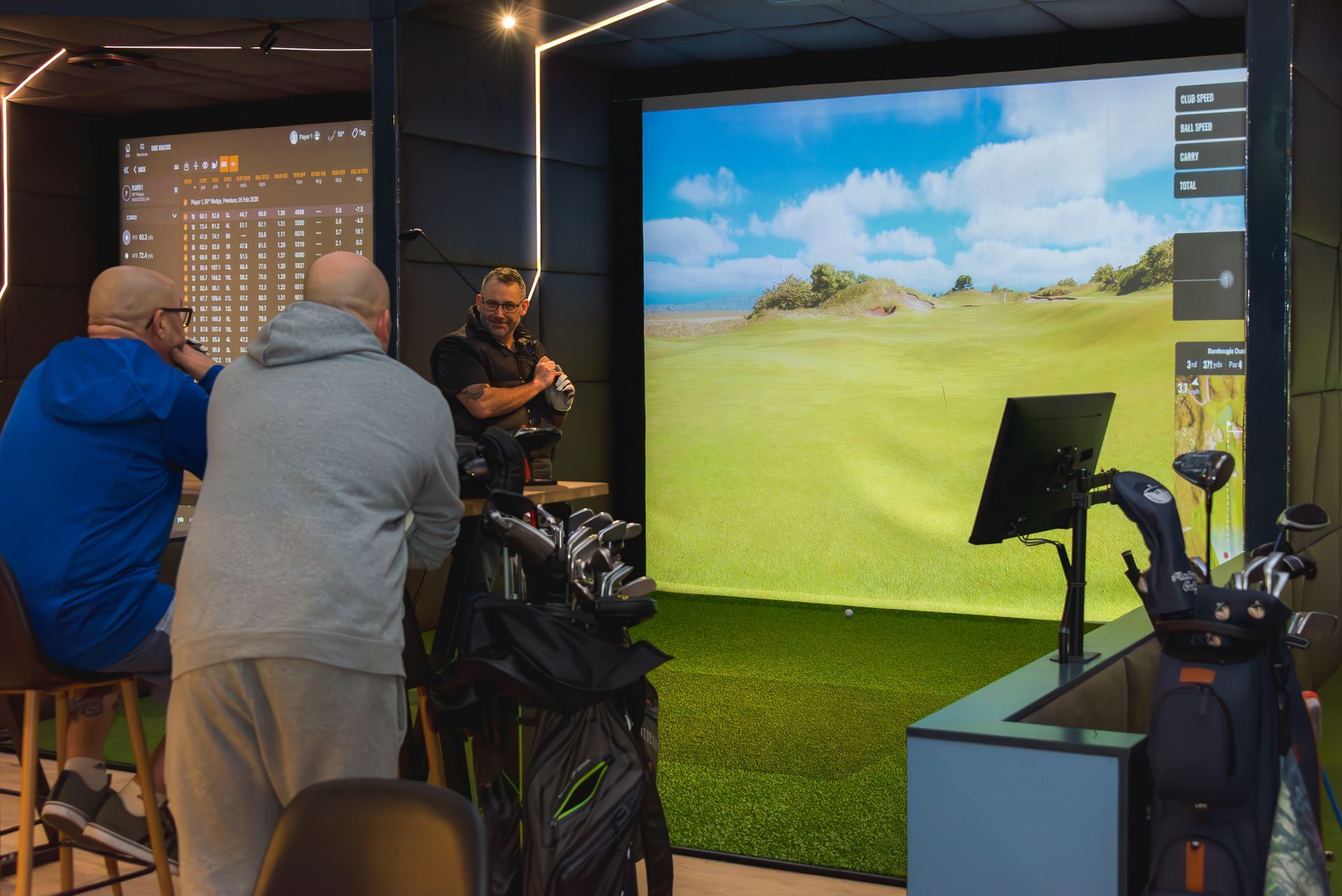 People playing a golf simulator indoors beside a large virtual course screen