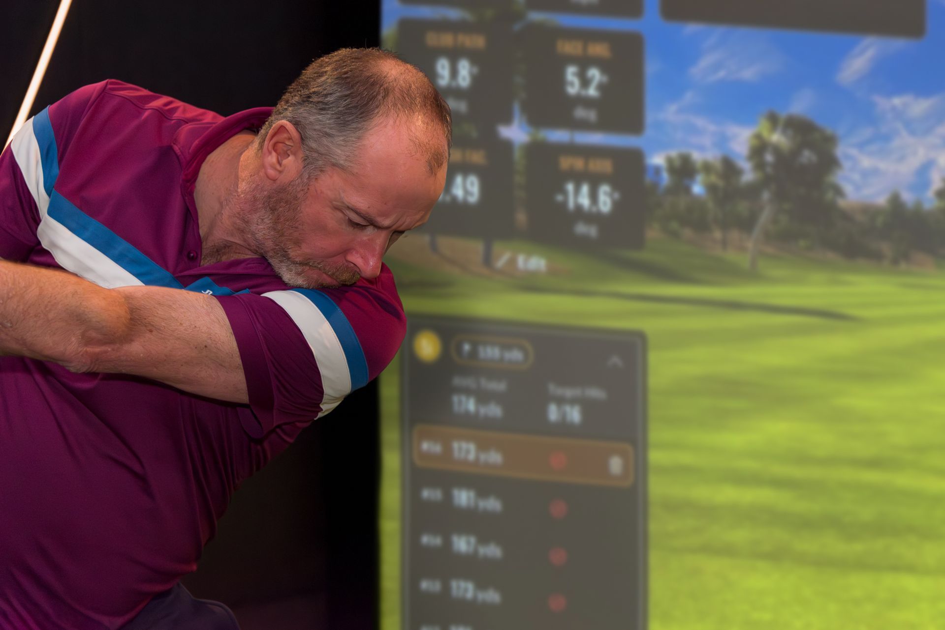 Golfer in a purple shirt swings in an indoor golf simulator with course graphics on screen