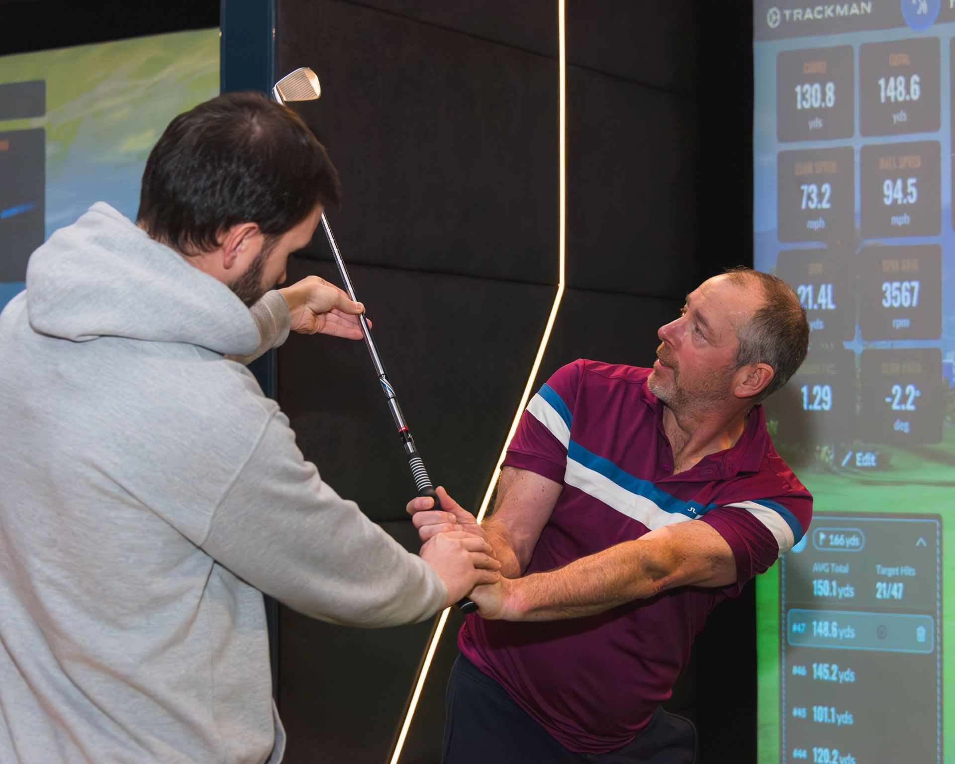 Two men indoors, one helping the other hold a golf club near a digital scoreboard.