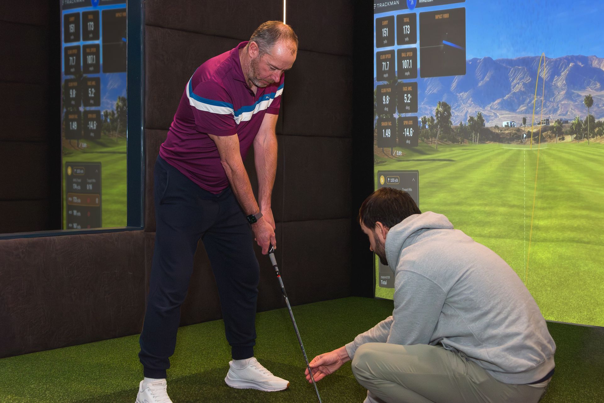 Two men practicing golf indoors with a simulator screen showing a green course and mountains.
