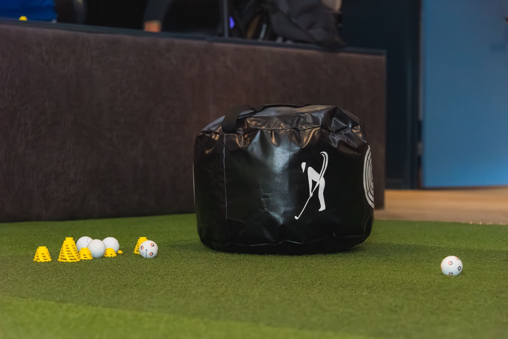 Golf driving range with a black ball bag and scattered yellow and white golf balls on green turf