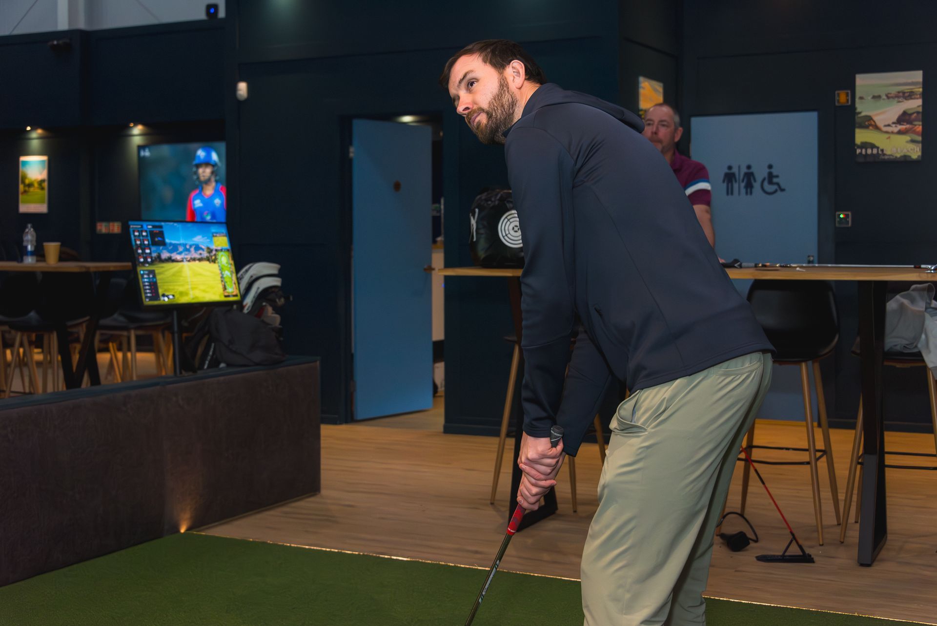 Man lining up a golf putt indoors on a green mat, holding a putter in a dimly lit room