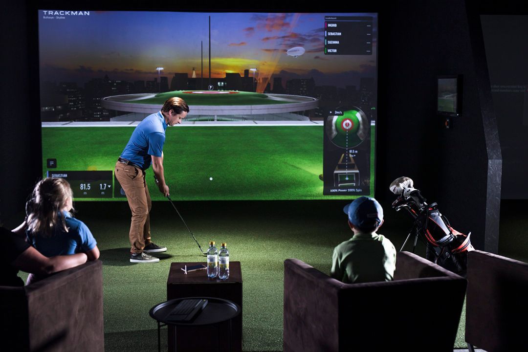 A man swings a golf club in a simulator, with two children watching on chairs in an indoor golf setting.